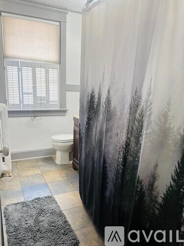 A bathroom with a toilet, a shower curtain with a forest scene on it, and a window with blinds.