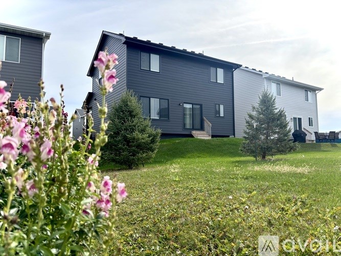 A house with a grey exterior is surrounded by green grass and pink flowers.