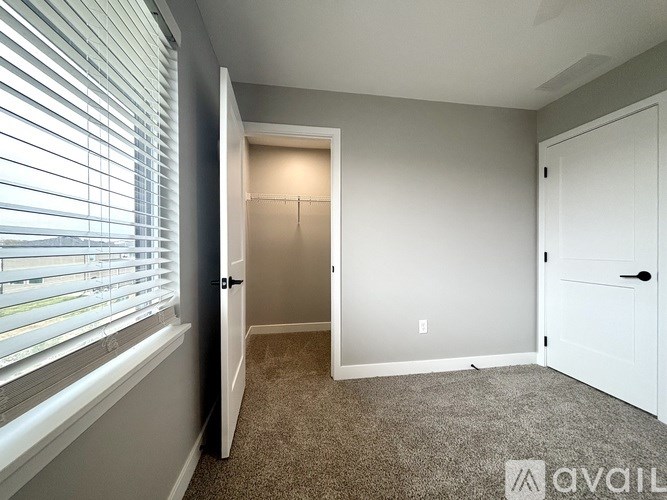 A room with a carpeted floor, a window with blinds, and a door leading to another room.