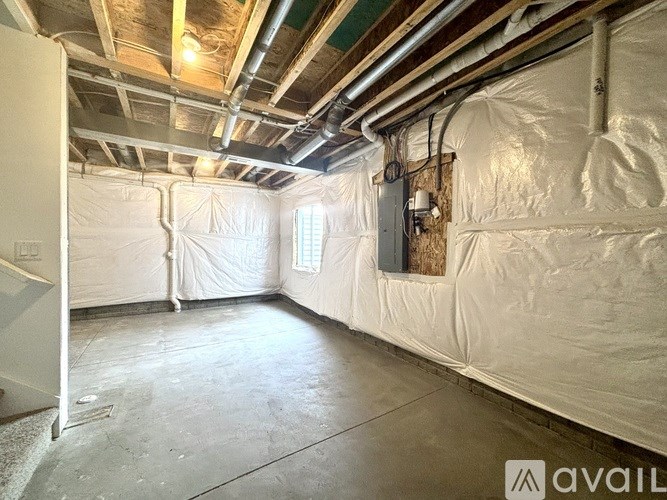A room with white walls and ceiling, covered in plastic sheeting, with visible pipes and ducts on the ceiling.