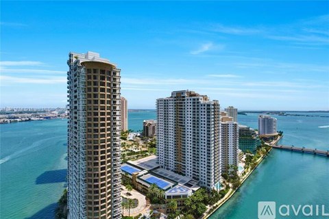 A tall building stands in front of a body of water with other buildings in the background.