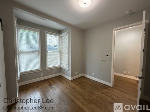 A room with wooden floors and white walls, featuring a window with blinds and a door.