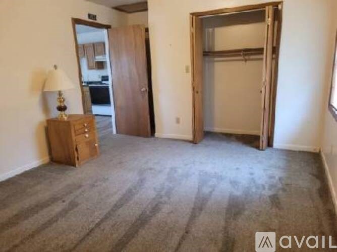 A room with a carpeted floor, a wooden dresser with a lamp, and a door leading to another room.