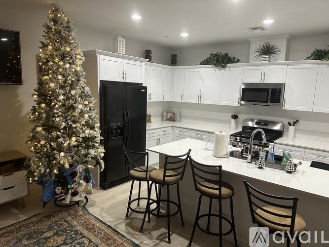 A Christmas tree stands in a kitchen with white cabinets and a black refrigerator.