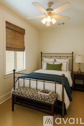 A bedroom with a bed, a window with a brown curtain, a ceiling fan, and a small table with a lamp.