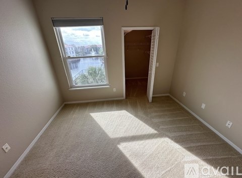 A room with a carpeted floor, a window with a view of a body of water and buildings, and a doorway leading to another room.