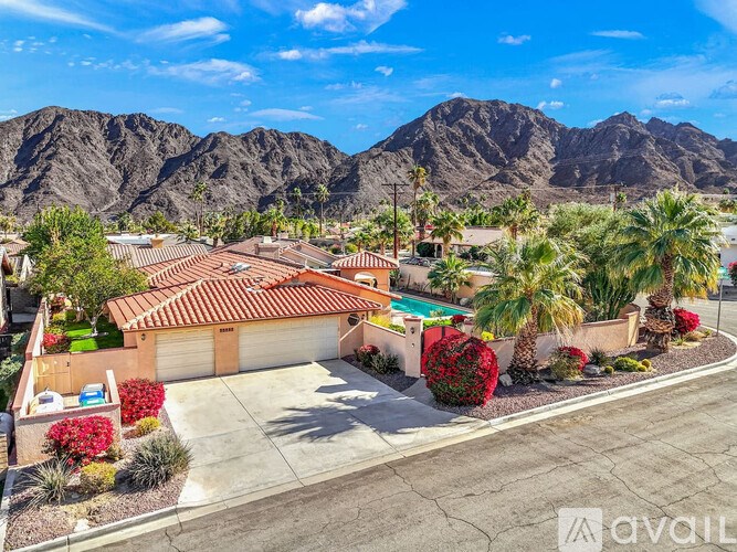 A house with a brown roof is surrounded by palm trees and red bushes.