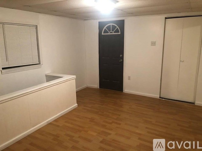 A room with wooden flooring, a window with blinds, a black cabinet with a mirror, and a white door.