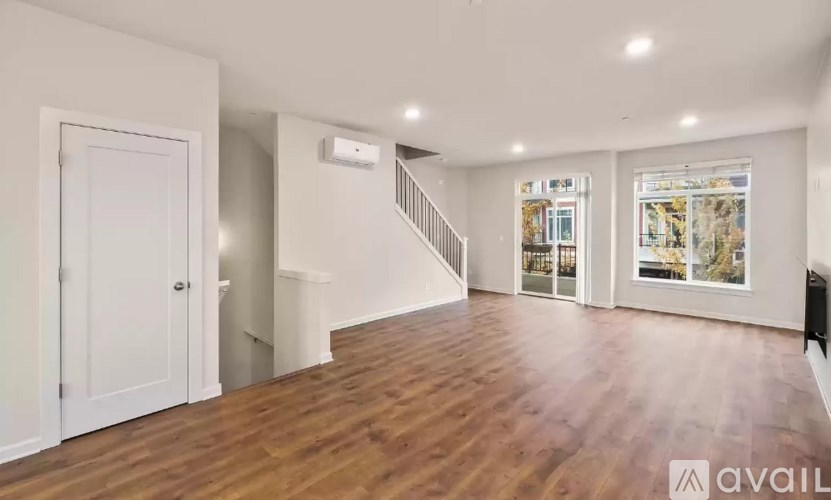 A spacious room with wooden floors and white walls, featuring a staircase and a large window with a view.