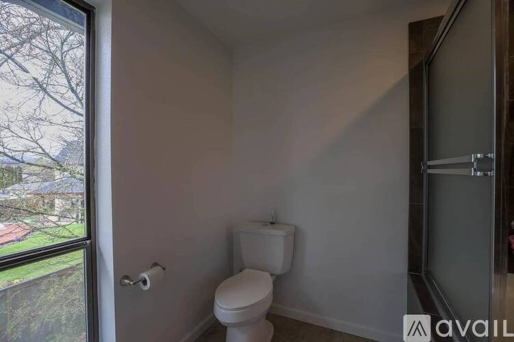 A white toilet sits in a small bathroom with a window overlooking a yard.