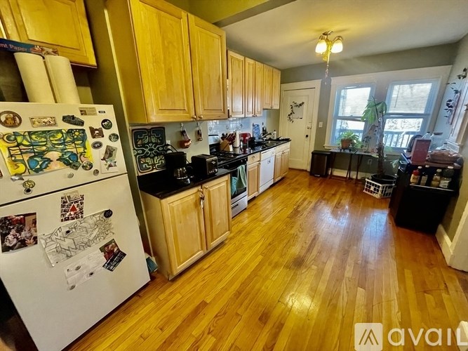 A kitchen with wooden floors and cabinets, a white fridge with magnets, and a window with a view of a tree.