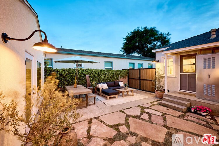 A patio with a table and chairs is surrounded by a stone floor and a hedge.