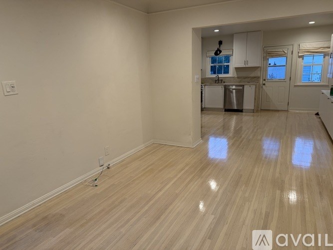 A room with wooden flooring and white walls, with a kitchen area visible in the background.