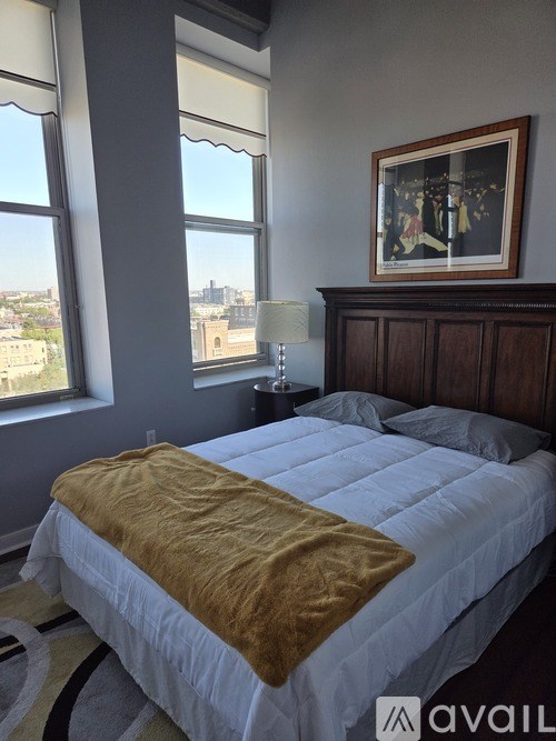 A bed with a white comforter and a gold blanket is in the foreground of a room with a framed picture on the wall.