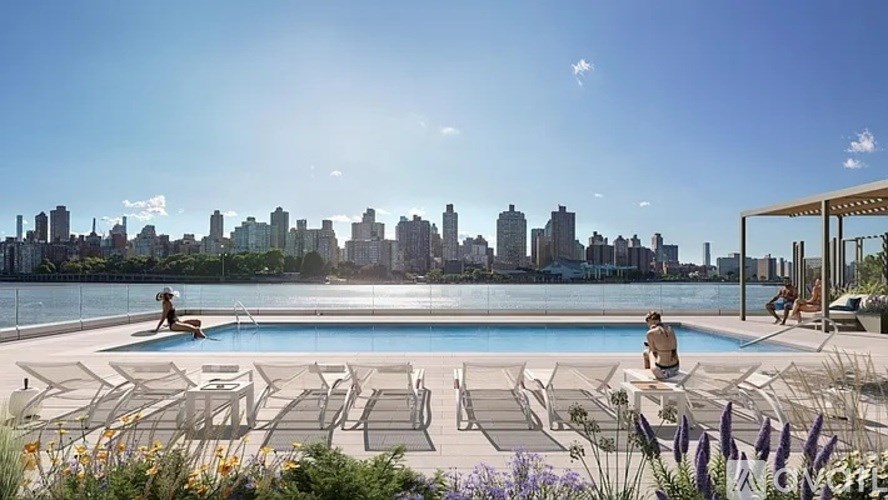 A pool with a city skyline in the background and a person sitting on a lounge chair.