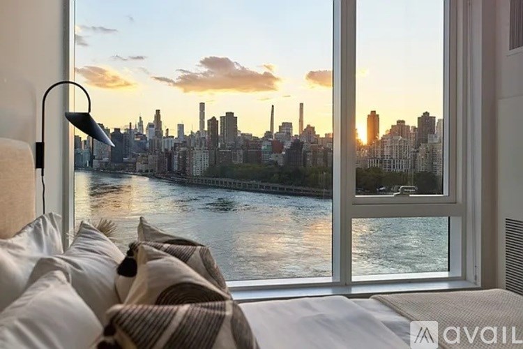 A bedroom with a large window overlooking a city skyline at sunset.