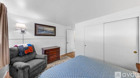 A bedroom with a grey sofa, a bed with a blue and white checkered cover, a wooden chest of drawers, a lamp, a picture on the wall, and a door.
