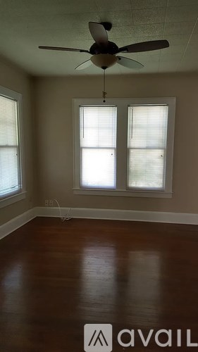 A room with a fan, two windows and wooden floors.