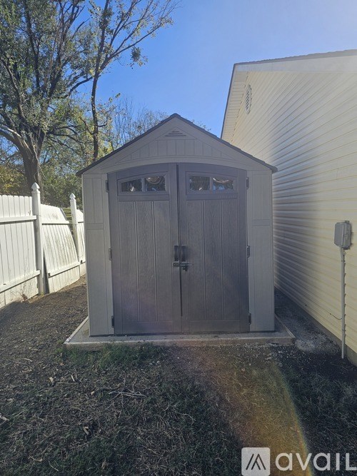 A small wooden shed with a brown door and windows is situated in a yard.