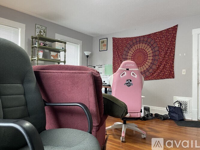 A pink office chair with a black logo on the back is in a room with a grey chair and a patterned wall hanging.