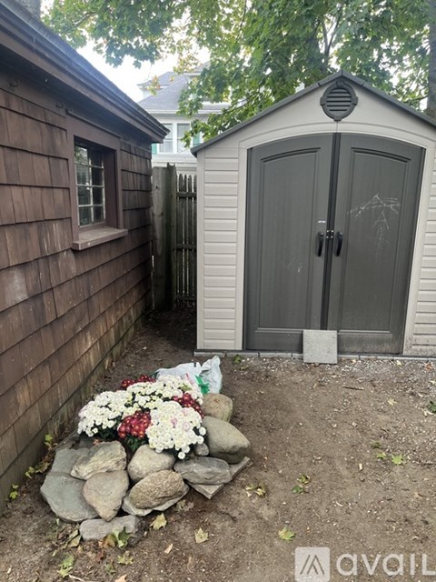 A small gray shed sits next to a wooden building with a stone planter in front of it.