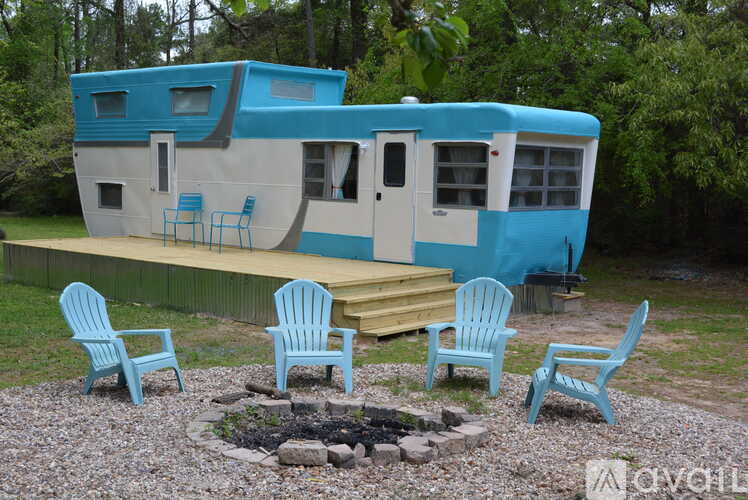 A blue and white trailer is parked in a gravel area with four chairs and a fire pit.