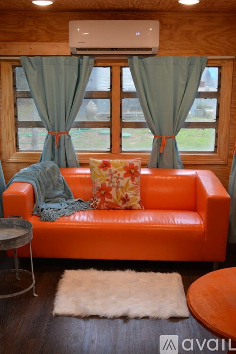 A bright orange couch with a floral pillow and a grey throw blanket sits in a room with a window.