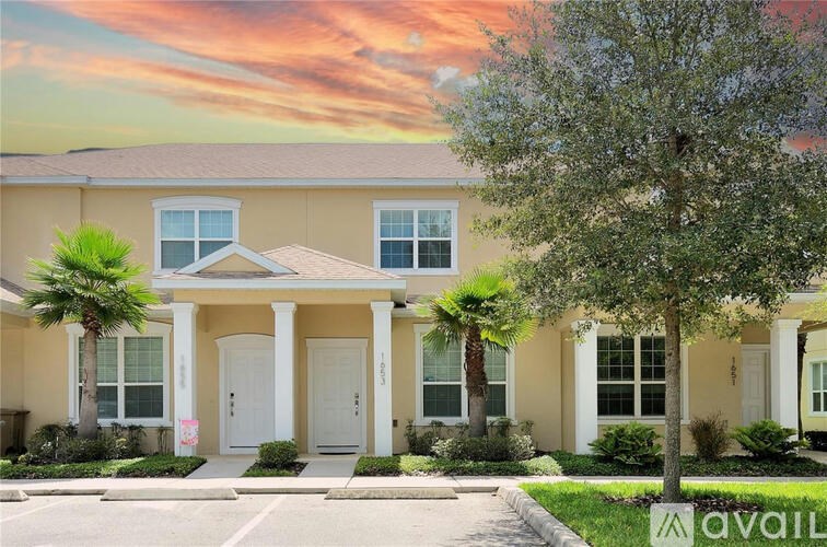 A house with a white door and windows is surrounded by greenery and has a tree in front of it.