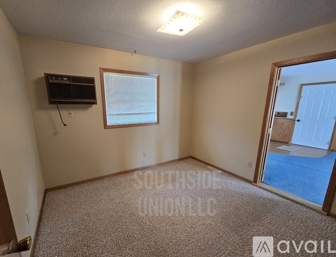 A room with a carpeted floor, a window with blinds, a flat-screen TV mounted on the wall, and a sliding glass door leading to a patio.