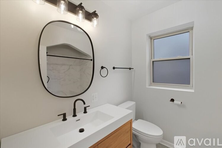 A bathroom with a round mirror, sink, toilet and window.