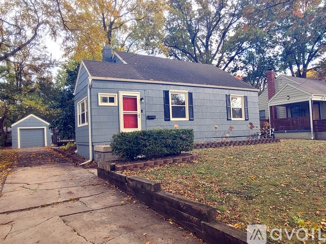 A small blue house with a red door is surrounded by trees and grass.