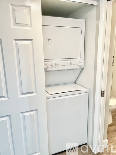 A white dryer is in a small space between a door and a toilet.