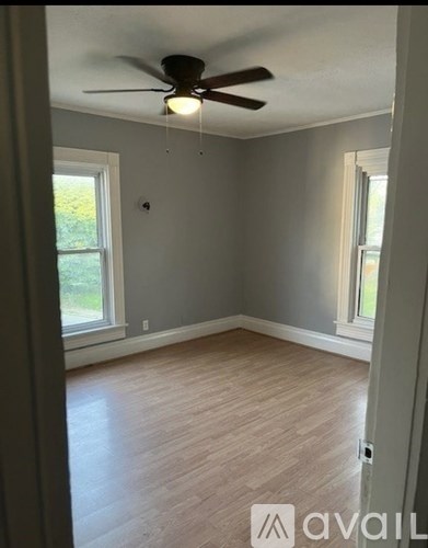A room with a ceiling fan and light fixture, wooden flooring, and two windows.