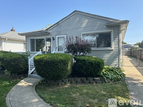 A house with a grey siding and a white door is surrounded by a well-kept lawn and a stone wall.