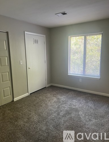 A room with a carpeted floor, a door, a window with blinds, and a wall-mounted light fixture.