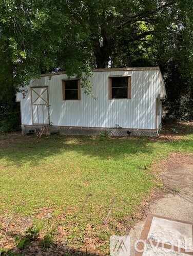 A white shed with a brown door and windows is surrounded by trees.