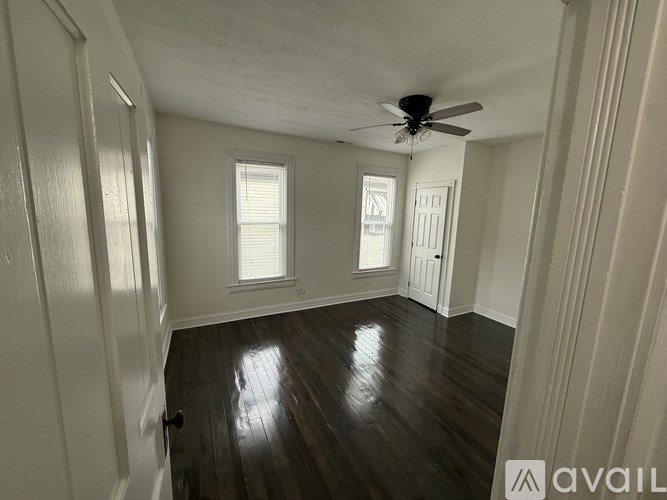 A room with wooden floors and white walls, featuring a ceiling fan and two windows.