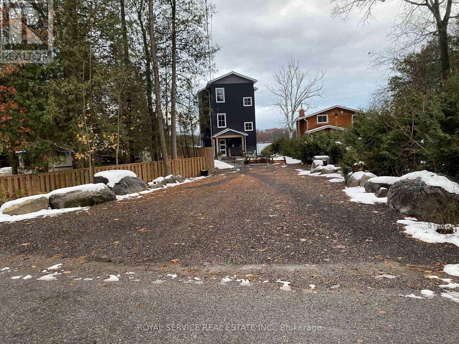 A gravel driveway leads to a large house with a fence and trees in the background.