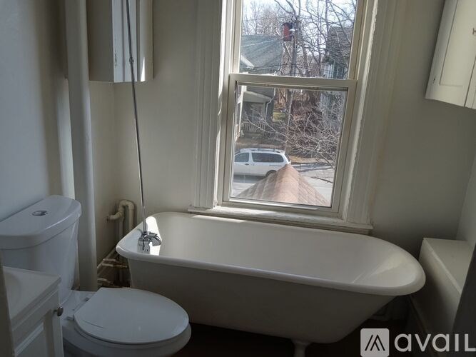 A white toilet sits next to a bathtub in a bathroom with a window overlooking a street.