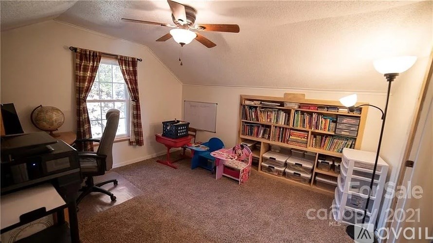 A room with a ceiling fan, a desk with a computer, a chair, a window with curtains, a bookshelf with books, and a lamp.