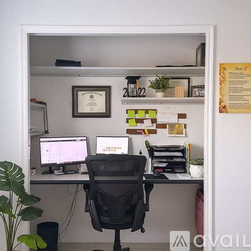 A black office chair is in front of a desk with a computer and a framed certificate on the wall.