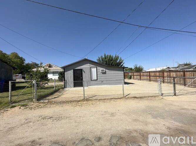 A small grey building with a black door is surrounded by a fence and a grassy area.