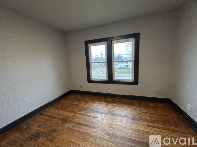 A room with wooden flooring and a window showing a view of a street with cars and a building.