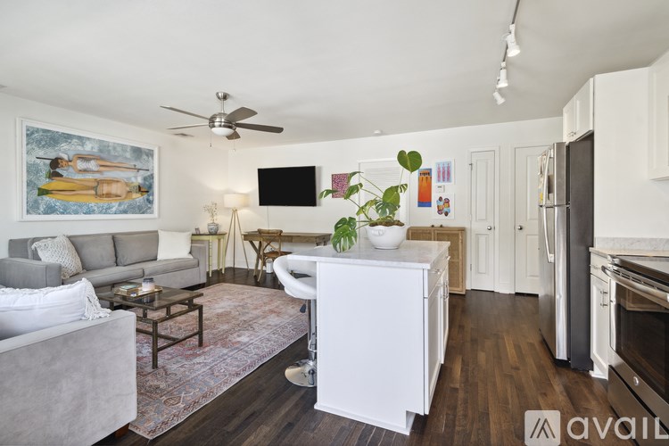 A living room with a grey couch, a white armchair, a coffee table, a painting, a TV, a ceiling fan, and a kitchen area with a refrigerator, oven, and cabinets.