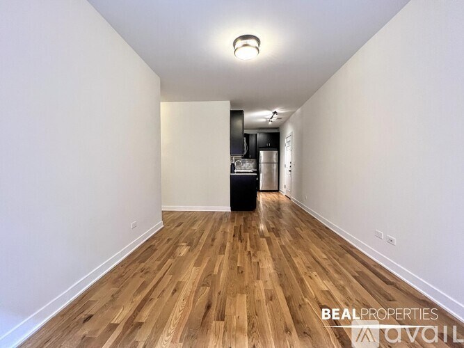 A room with wooden flooring and white walls, with a kitchen area visible in the background.