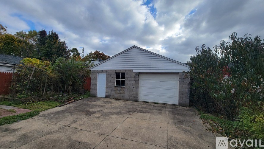 A detached garage with a white door and a grey roof is surrounded by greenery and a cloudy sky.