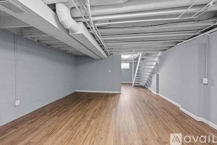 A spacious room with wooden flooring and white walls, featuring a staircase and a ceiling with exposed pipes.