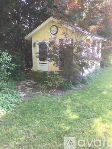 A small yellow building with a clock on the front is surrounded by greenery.