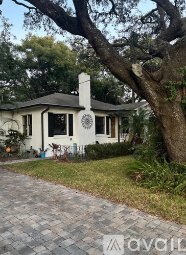 A white house with a black roof and a chimney is surrounded by greenery.
