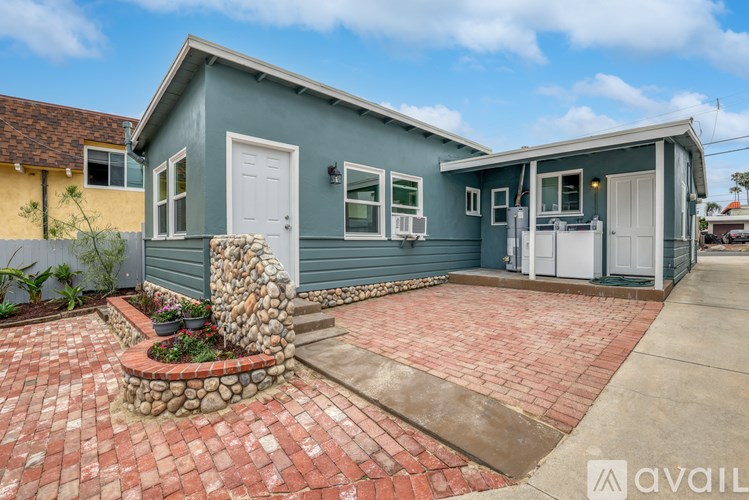 A blue house with a white door and windows is surrounded by a brick walkway and a stone planter.